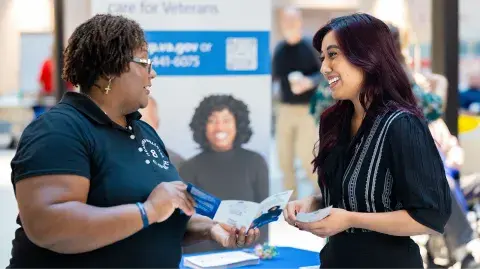 Two young women of color talk with each other while examining a Million Veteran Program pamphlet. They are standing next to a table and an MVP pull up banner.